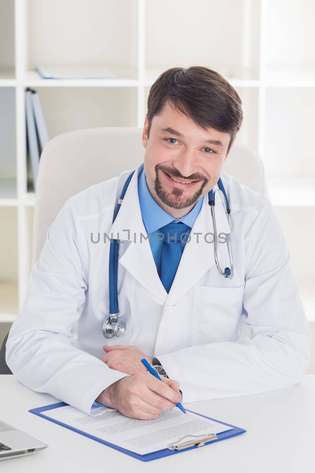 Male doctor working at office desk and smiling at camera , office interior on background