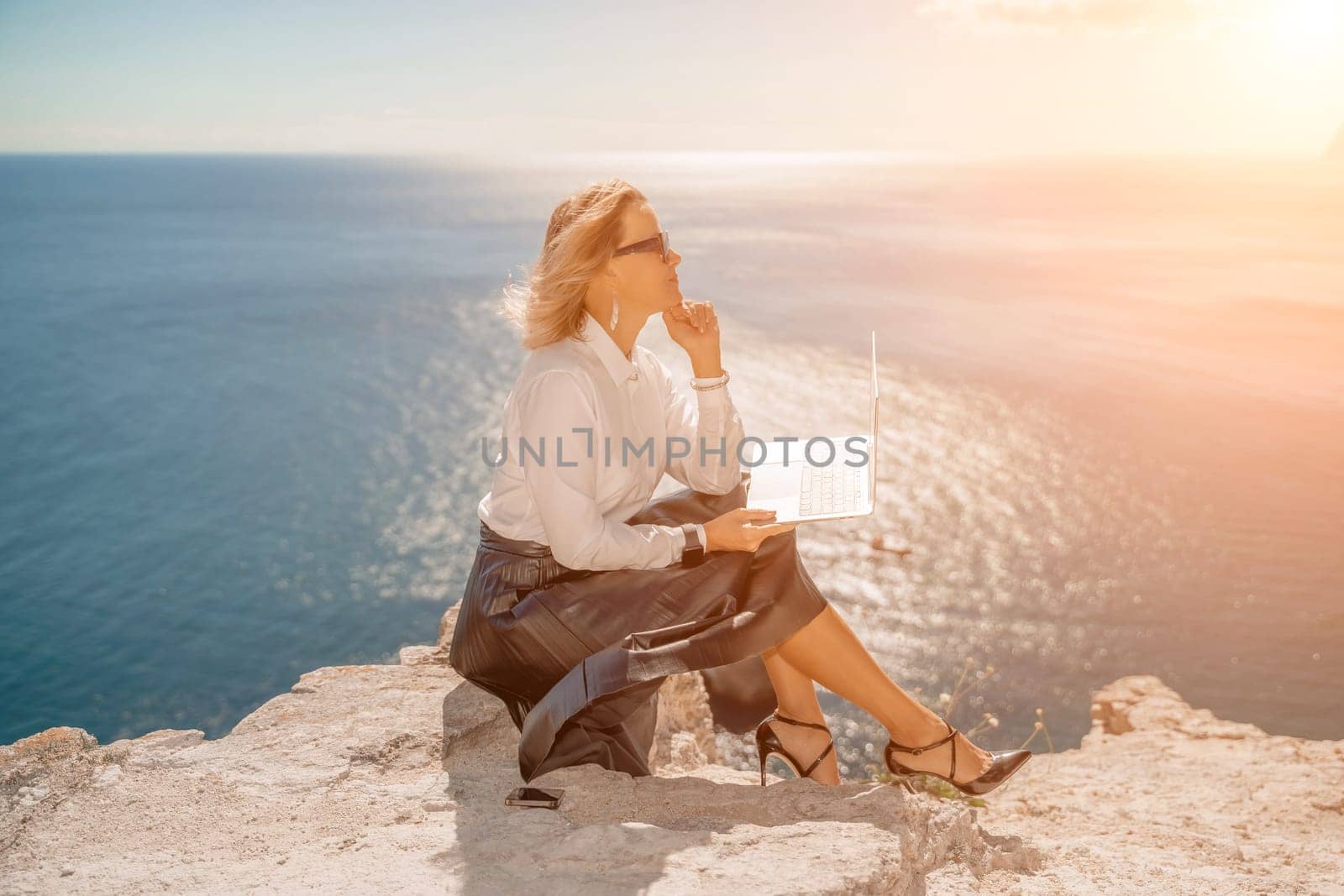 Business woman on nature in white shirt and black skirt. She works with an iPad in the open air with a beautiful view of the sea. The concept of remote work