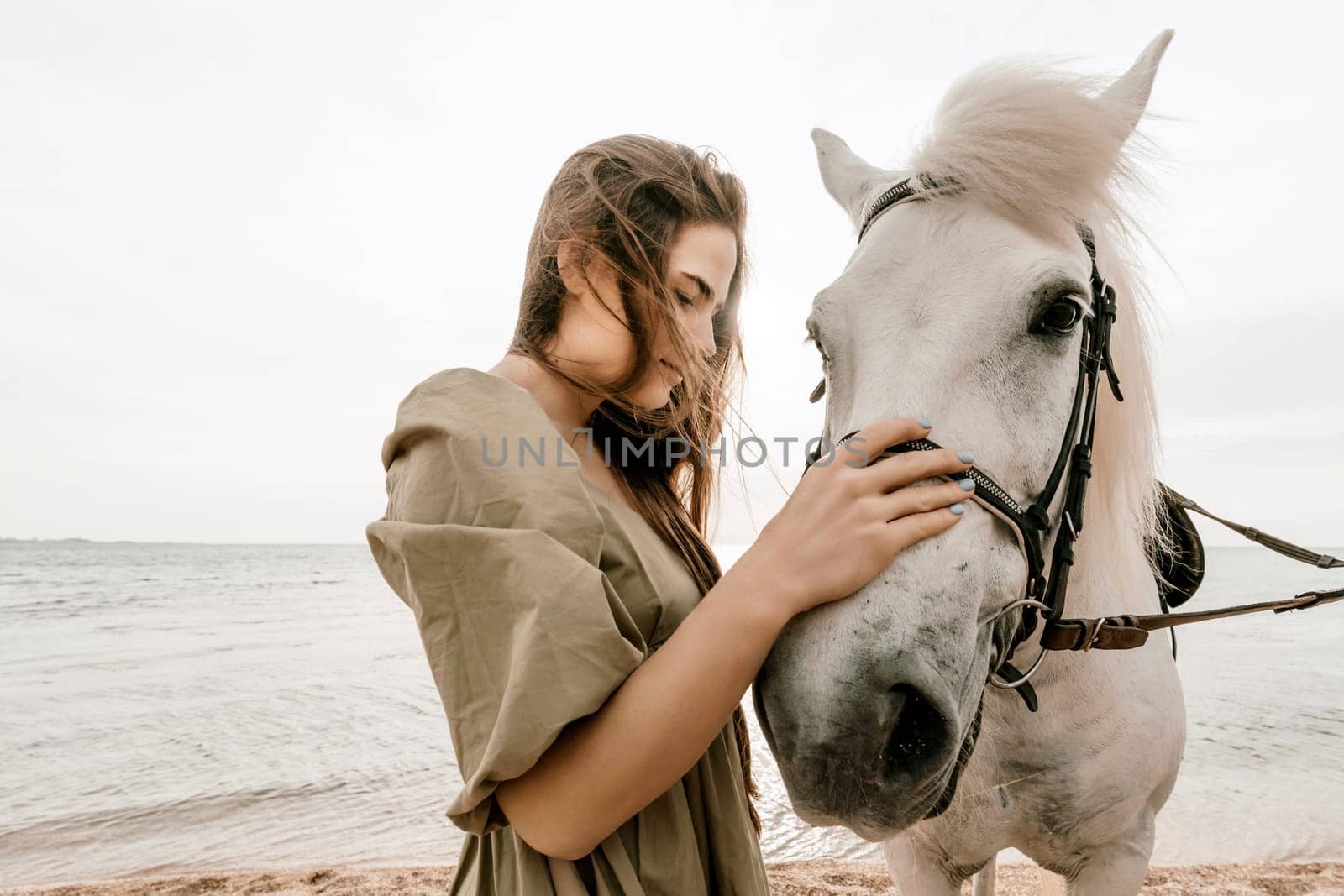 A woman in a dress stands next to a white horse on a beach, with the blue sky and sea in the background