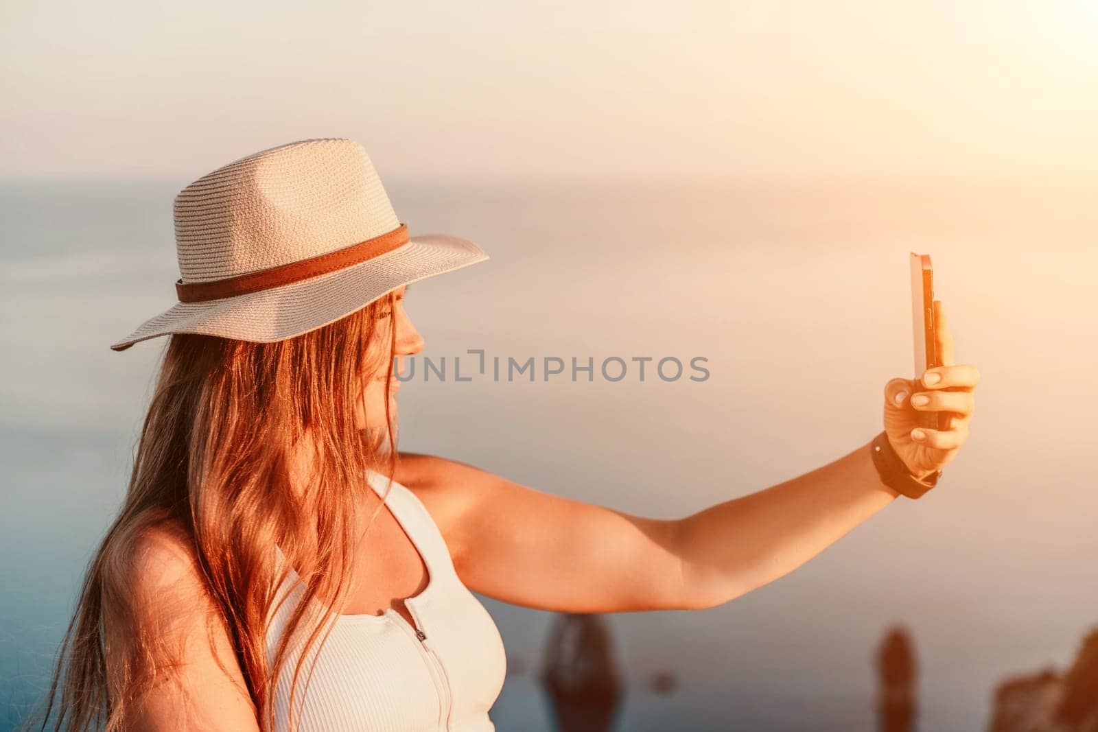Selfie woman in a hat, white tank top, and shorts captures a selfie shot with her mobile phone against the backdrop of a serene beach and blue sea. by Matiunina