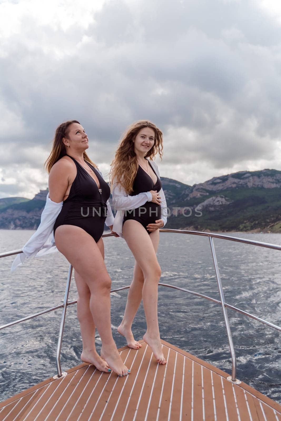 Pregnant on a yacht. Happy models in a swimsuit posing on a yacht against sky with clouds and mountains