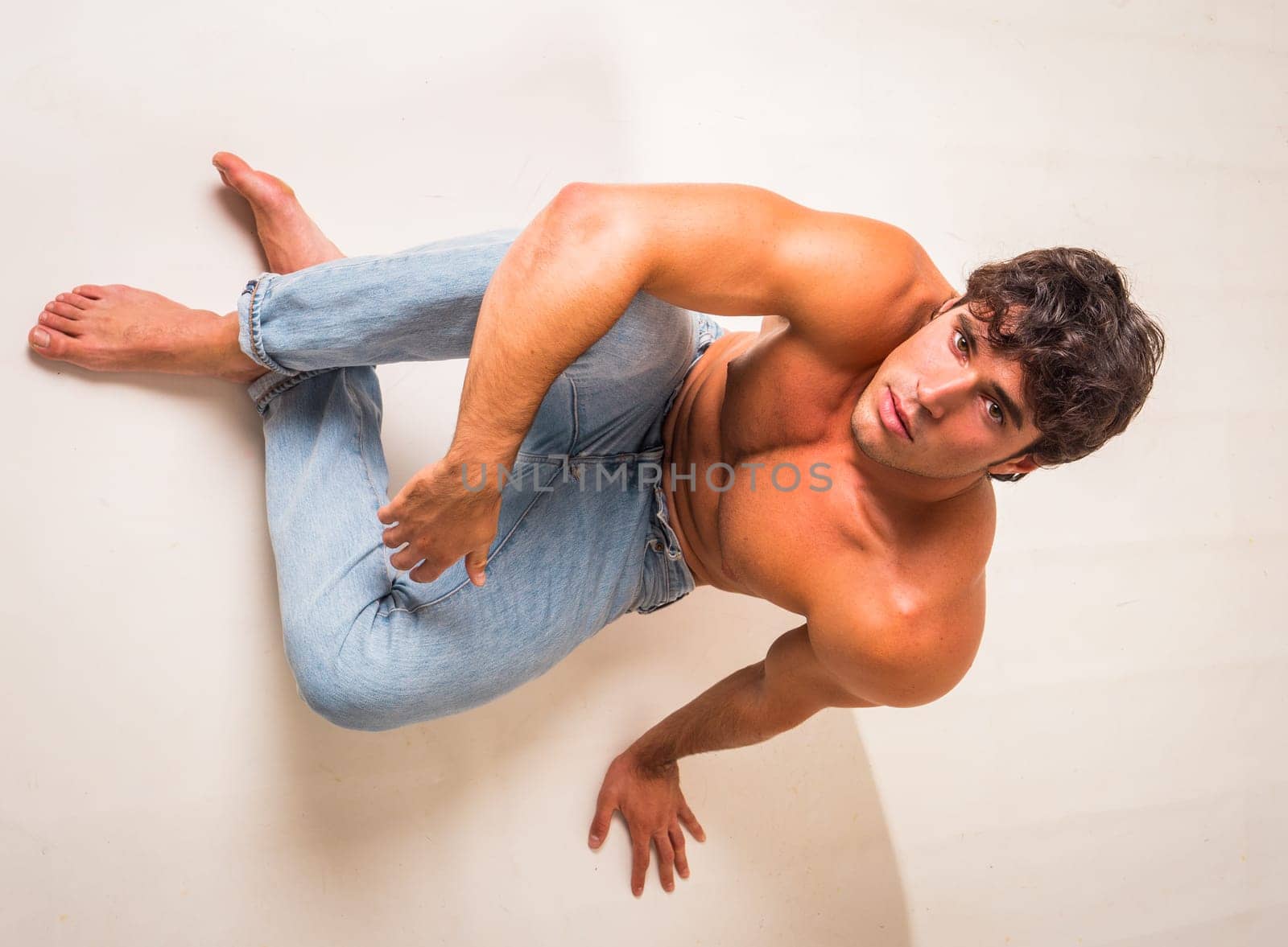 Young handsome shirtless man sitting on floor in studio against white, looking up