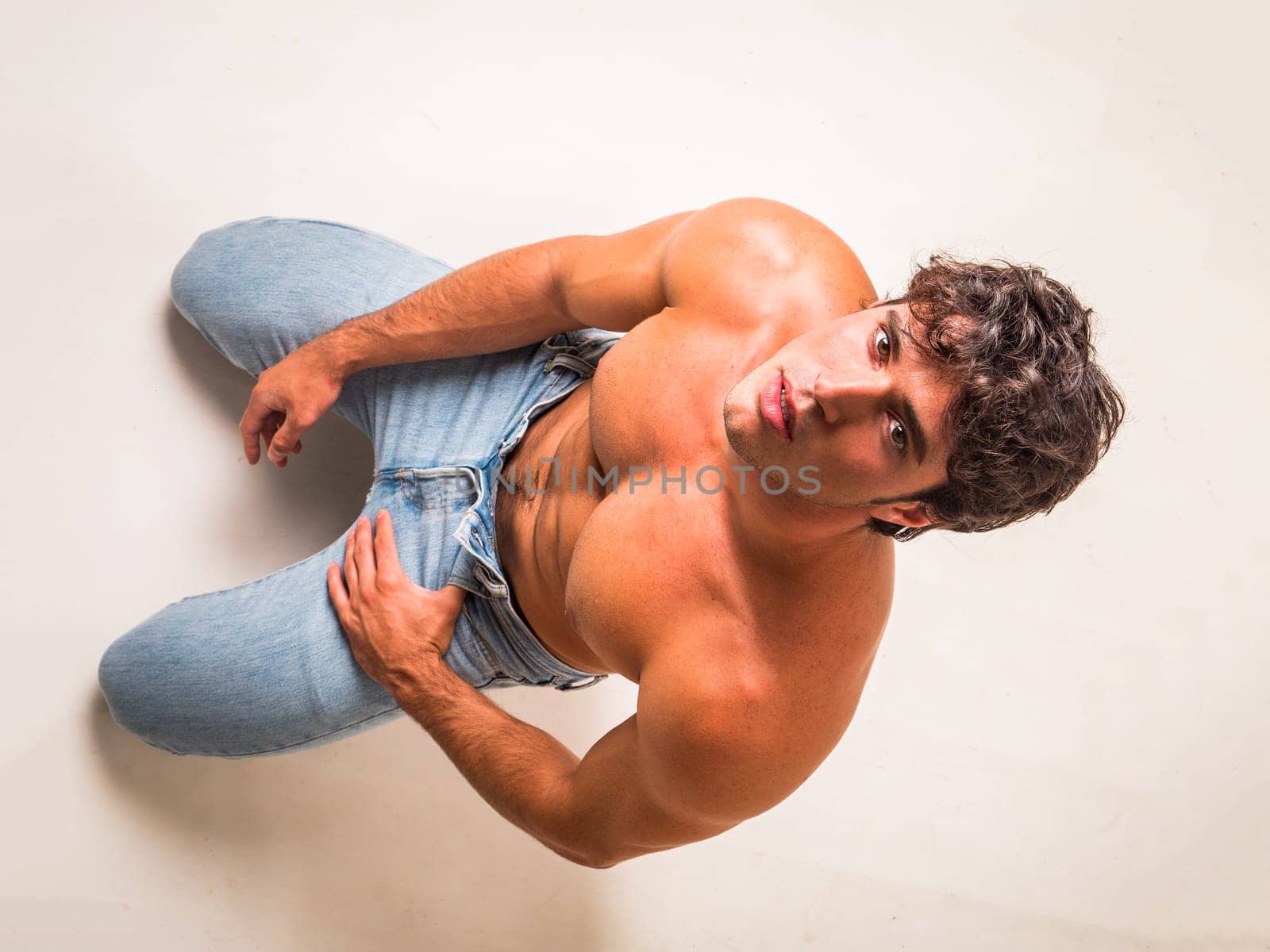 Young handsome shirtless man sitting on floor in studio against white, looking up