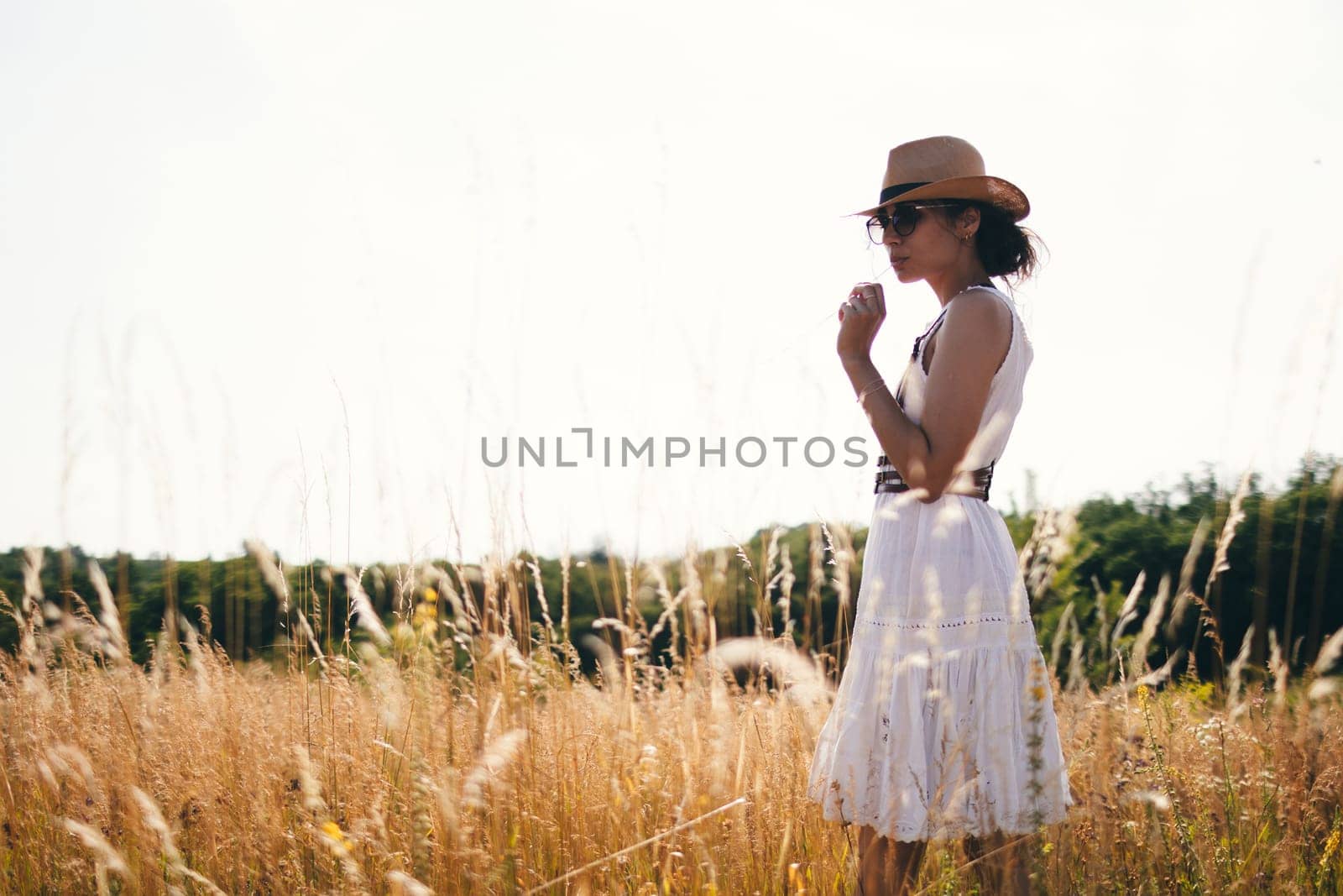 Portrait of young woman walking among high grasses in summer meadow wearing straw hat and linen dress enjoying nature. Harmony and balance by sarymsakov