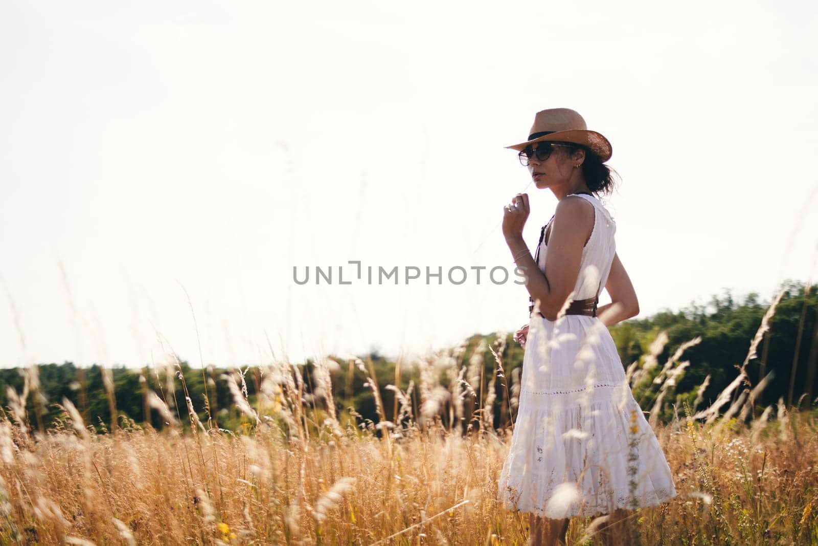 Portrait of young woman walking among high grasses in summer meadow wearing straw hat and linen dress enjoying nature. Harmony and balance.