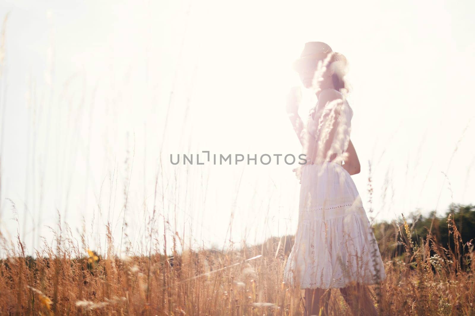 Portrait of young woman walking among high grasses in summer meadow wearing straw hat and linen dress enjoying nature. Harmony and balance by sarymsakov