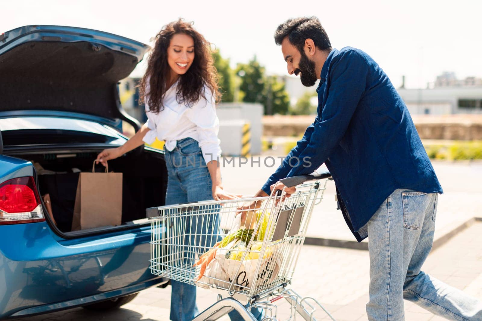 Family shopping day. Happy arab couple putting bags full of fresh food in car, standing outdoors after successful shopping in mall on weekend
