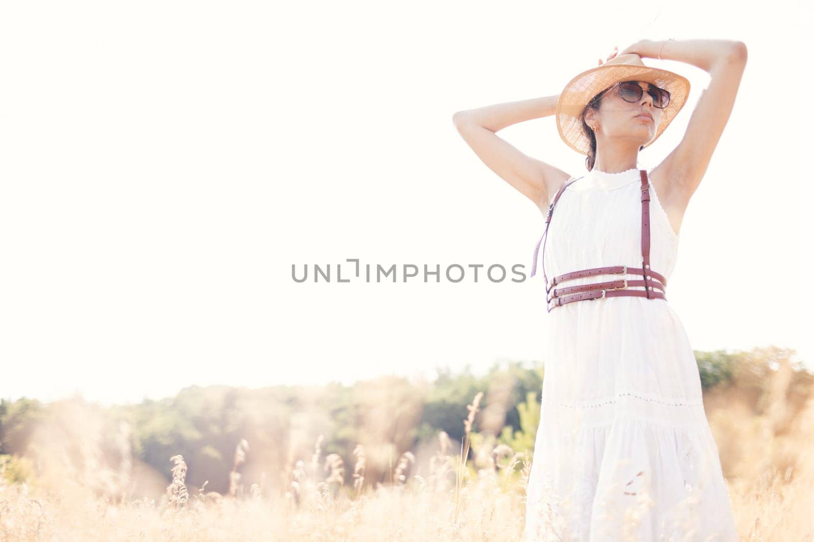 Spirit of freedom. An attractive boho girl in blouse, hat and sunglasses standing on the field on the background of a blue sky. Summer vacation, traveling. Bohemian, modern hippie style