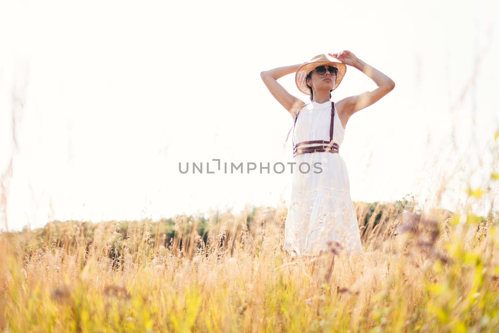 Spirit of freedom. An attractive boho girl in blouse, hat and sunglasses standing on the field on the background of a blue sky. Summer vacation, traveling. Bohemian, modern hippie style