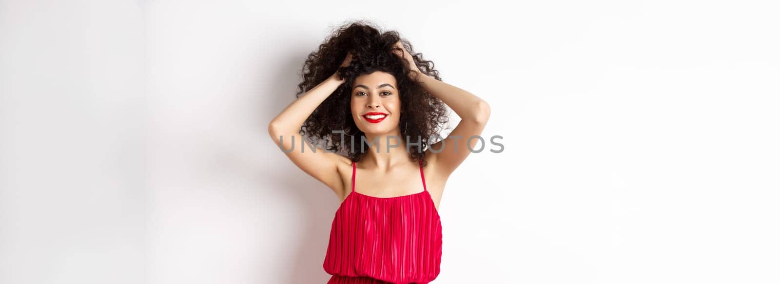 Beauty and fashion. Carefree woman in red dress and makeup, touching curly hair and smiling happy, standing on white background.