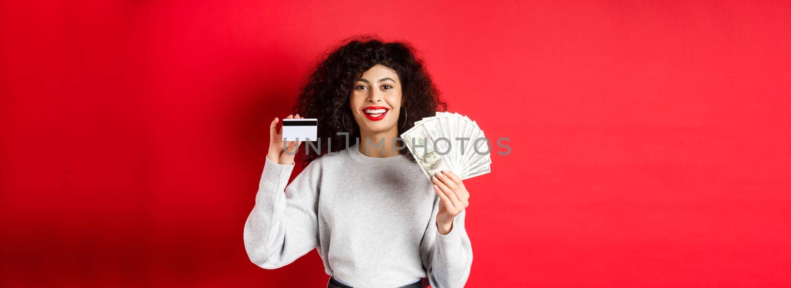 Portrait of stylish young woman with curly hair, showing money in cash and plastic credit card, red background.