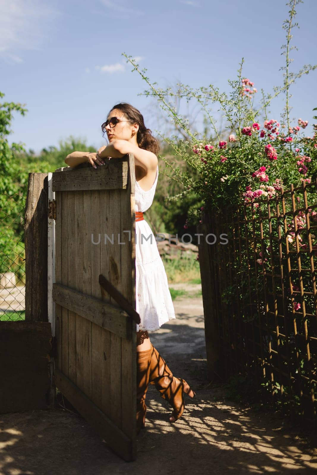 Young fashionable woman stands near fence in the garden or backyard of countryside house. Summer time, routine, relax, harvest concept.