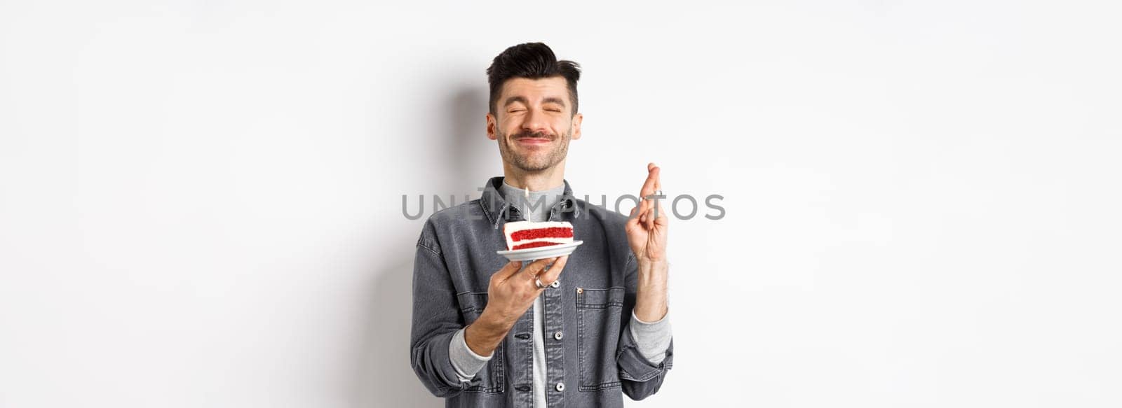 Hopeful birthday guy making wish with fingers crossed, holding bday cake on party, standing against white background.