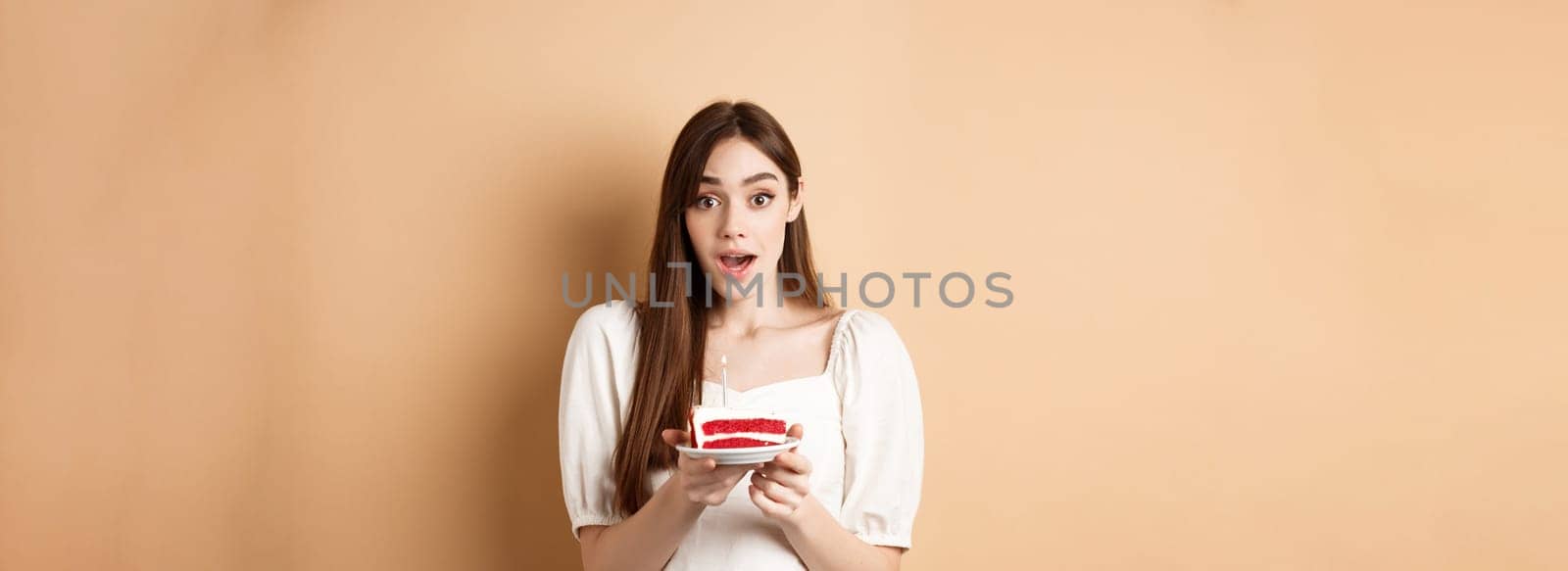 Excited birthday girl looking amazed, holding cake with candle, making b-day wish, standing on beige background.