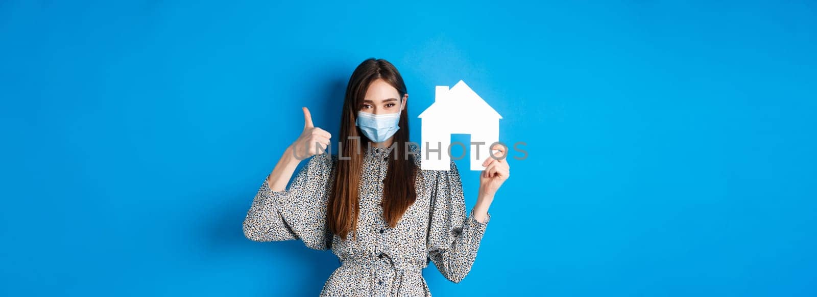 Real estate, covid-19 and pandemic concept. Candid woman in medical mask showing thumb up and paper house cutout, reanting apartment, standing on blue background.