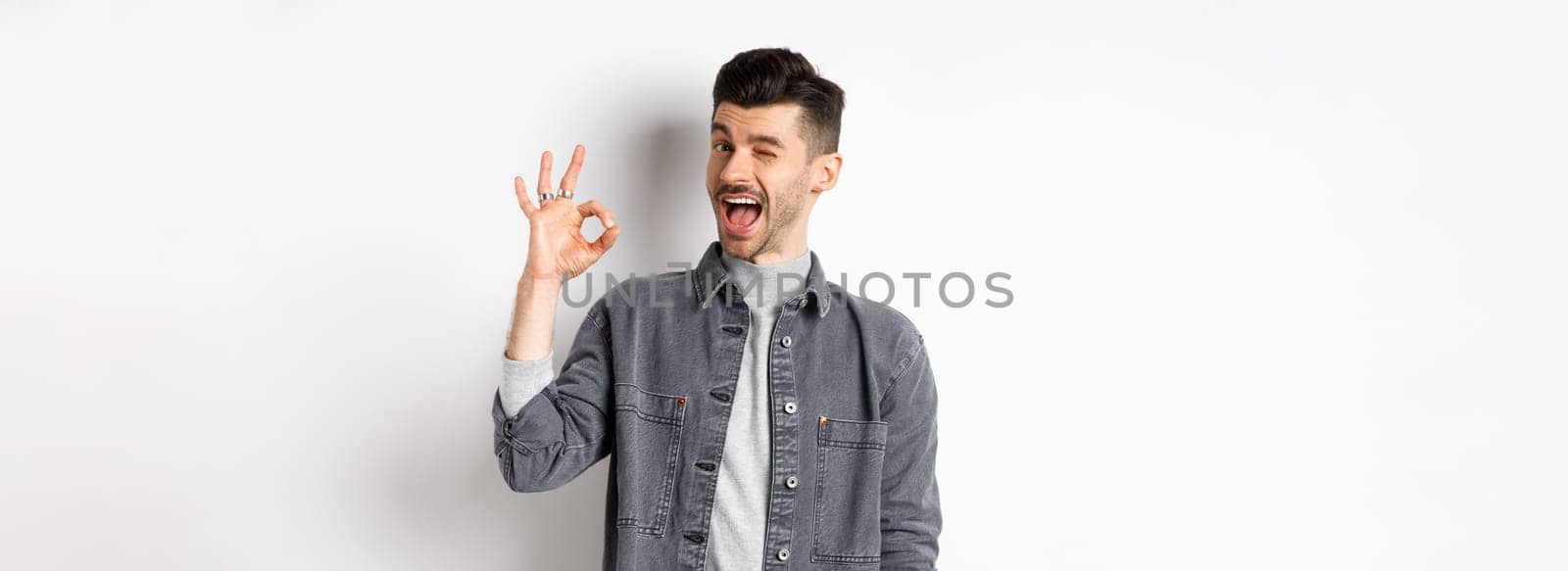 Excited handsome guy with moustache, winking and showing okay sign, smiling pleased, assure all good, praise nice work, making good job gesture, standing against white background.