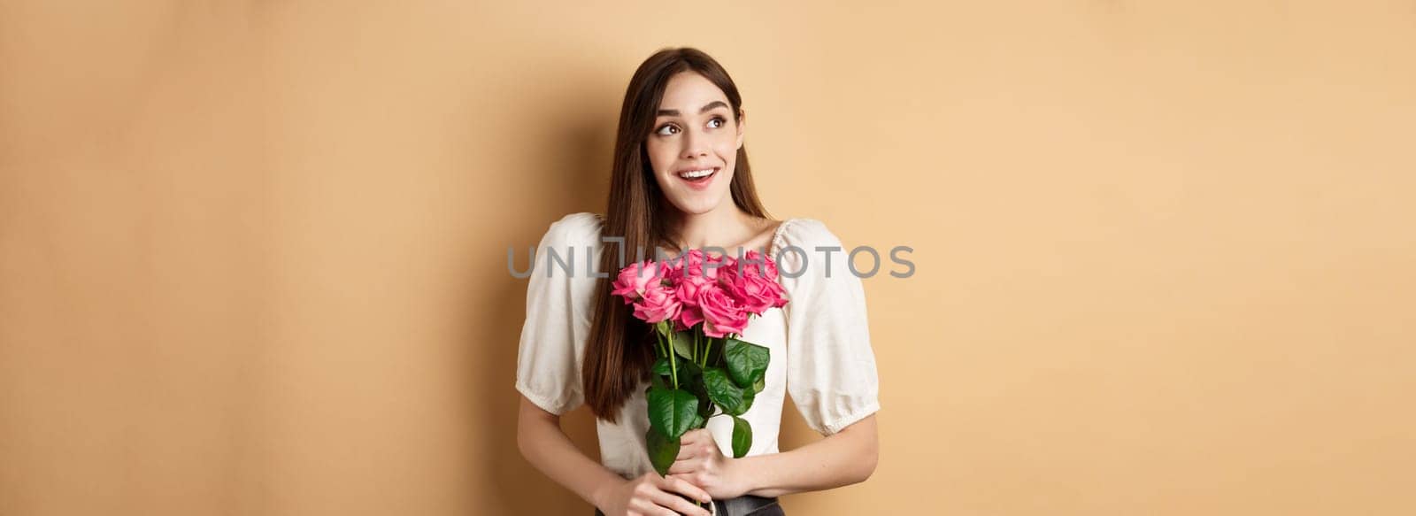 Happy valentines day. Dreamy smiling woman receive bouquet of pink roses and looking aside with love, standing on beige background.
