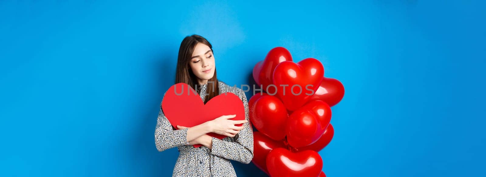 Valentines day. Romantic pretty woman in dress hugging big red heart cutout and looking dreamy, thinking of love, standing on blue background.
