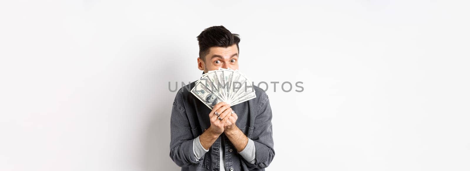Excited funny guy hiding face behind dollar bills and smiling, showing money in cash, standing against white background.