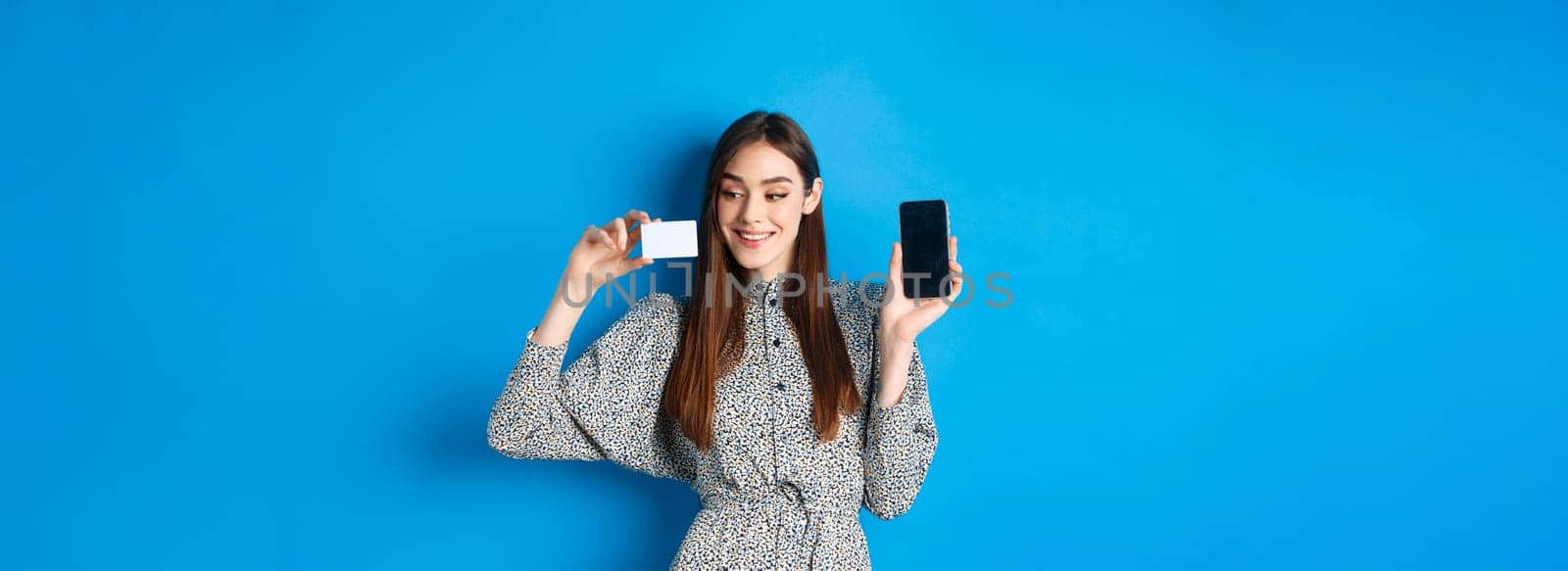 Online shopping. Beautiful female model showing empty cellphone screen and plastic credit card, smiling pleased, buying in internet store, blue background.