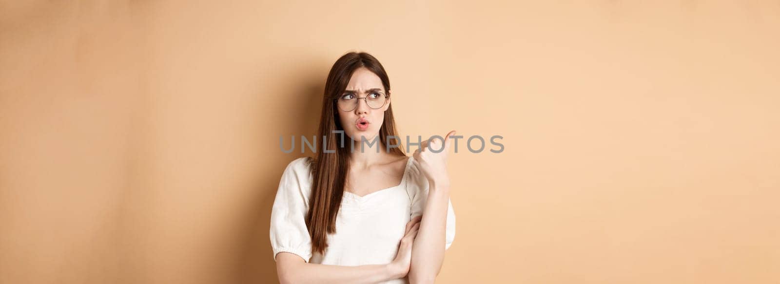 Confused and suspicious young woman in glasses frowning, pointing and looking aside with displeased face expression, standing on beige background by Benzoix