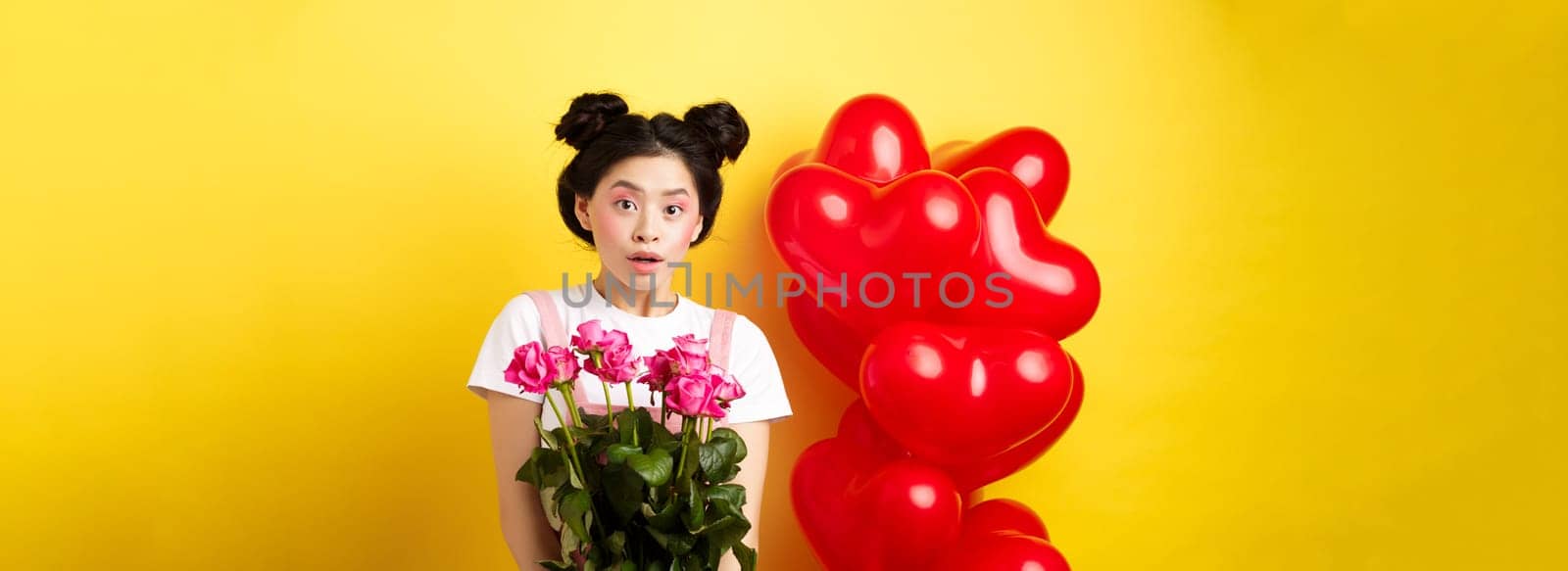 Happy Valentines day. Surprised asian girl receive bouquet of pink roses from lover, looking with awe at camera, standing near red heart balloons, yellow background by Benzoix