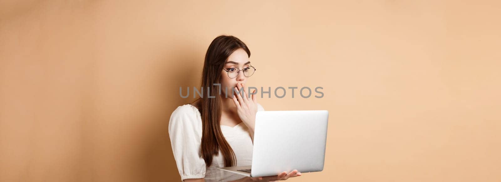 Shocked girl look at laptop screen, covering mouth with amazed face, standing in glasses on beige background by Benzoix