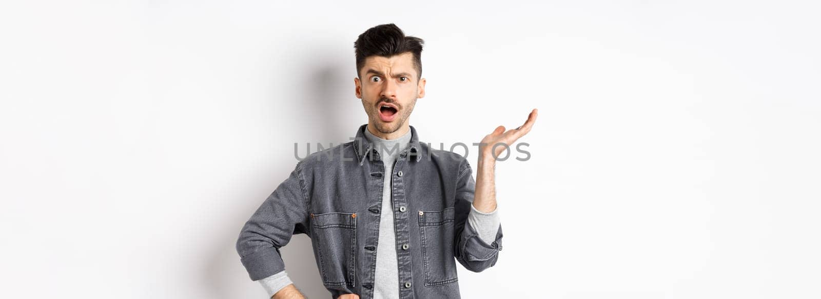 Confused and annoyed guy raising hand up and complaining, asking wtf happened, cant understand, standing on white background.