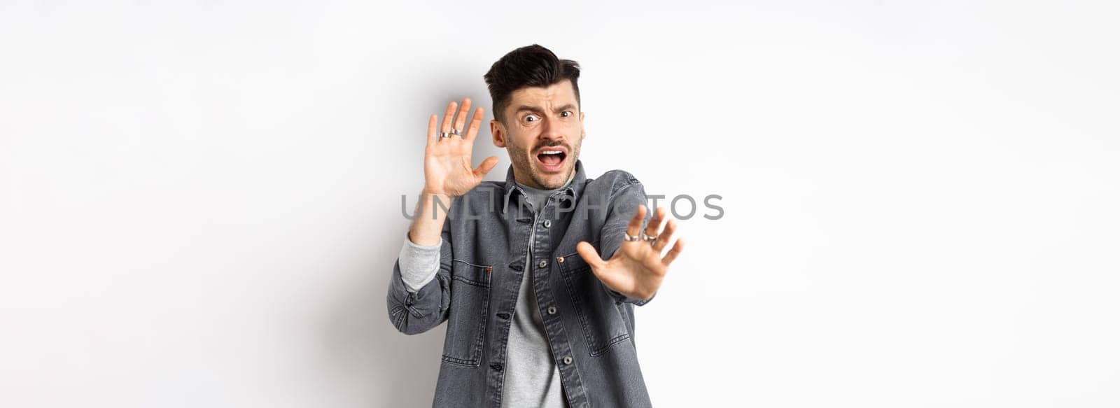 Scared timid guy screaming and raising hands in pleading pose, begging to stop, defending from assault, standing on white background.