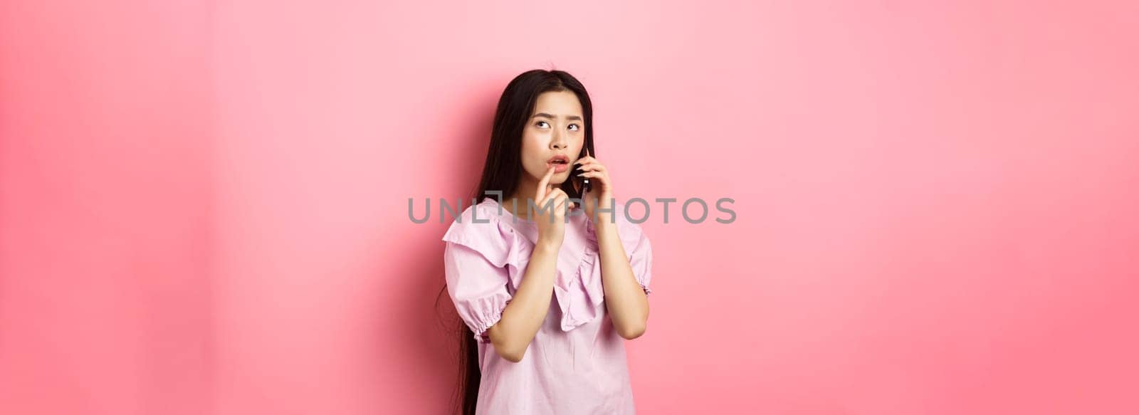 Thoughtful asian woman talking on mobile phone and thinking, touching lip and look up pensive, standing against pink background.