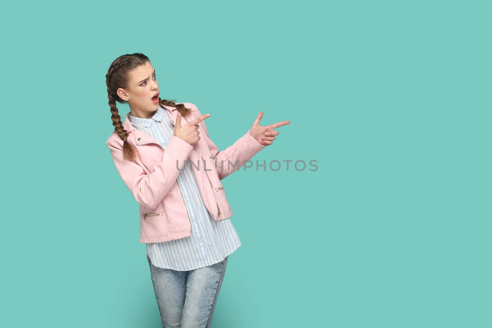 Portrait of shocked astonished teenager girl with braids wearing pink jacket pointing aside at advertisement area, space for promotion. Indoor studio shot isolated on green background.