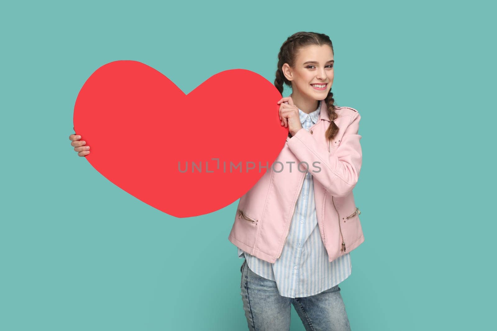 Portrait of extremely joyful cheerful teenager girl with braids wearing pink jacket, showing big red heart, smiling to camera. Indoor studio shot isolated on green background.