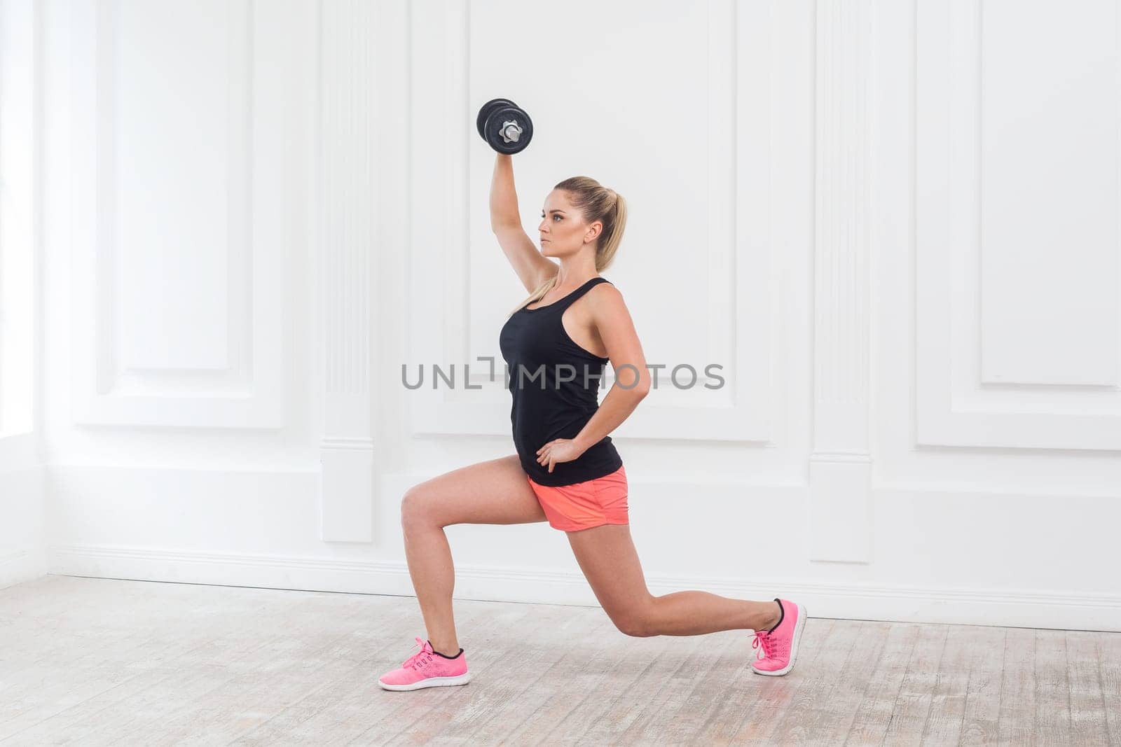 Side view portrait of young athletic beautiful blonde woman wearing pink shorts and black top holding dumbbell, doing exercise for arms at the gym on white wall. Indoor studio shot.