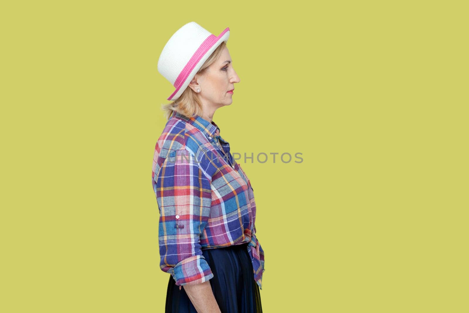 Side view portrait of serious strict mature woman wearing checkered shirt and hat standing looking ahead, being in bad mood. Indoor studio shot isolated on yellow background.