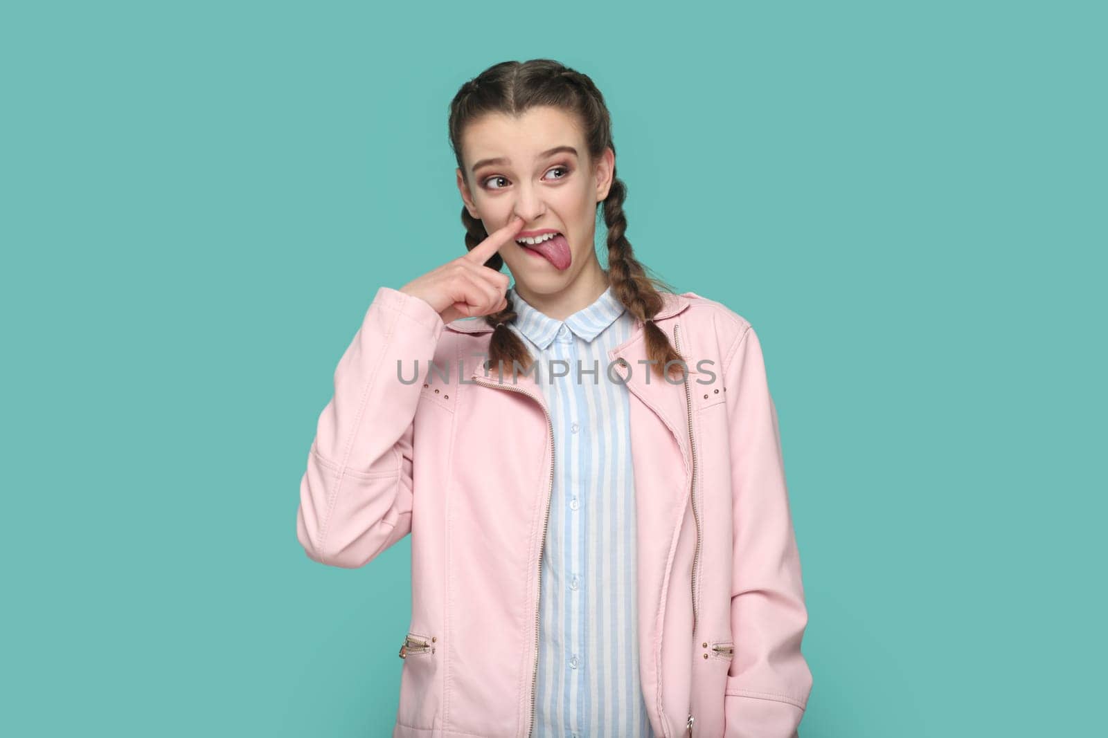 Portrait of funny childish teenager girl with braids wearing pink jacket standing with finger in nose, sticking tongue out, looking away. Indoor studio shot isolated on green background.