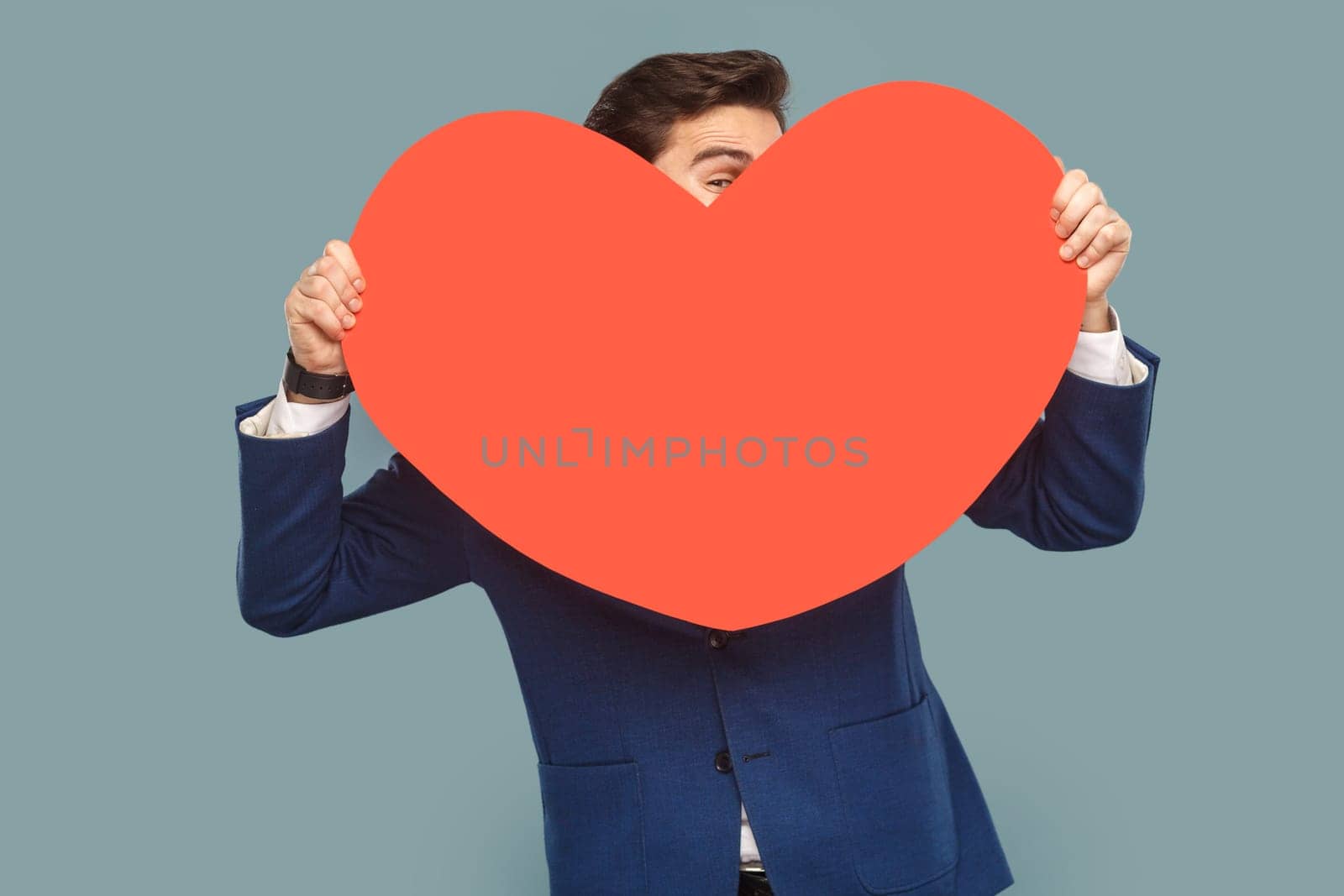 Portrait of funny optimistic shy man standing peeking out of big red heart, expressing love, being shy, wearing white shirt and jacket. Indoor studio shot isolated on light blue background.