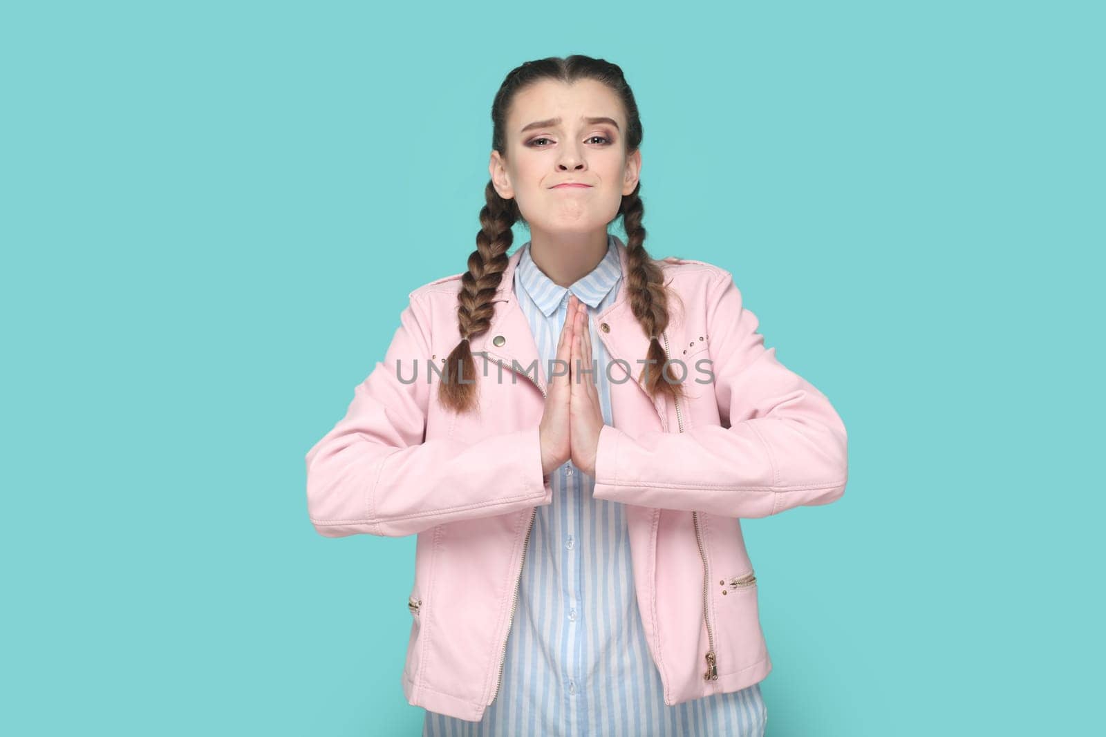Portrait of hopeful teenager girl with braids wearing pink jacket standing keeps hand in praying gesture, pleading, asking to forgive. Indoor studio shot isolated on green background.