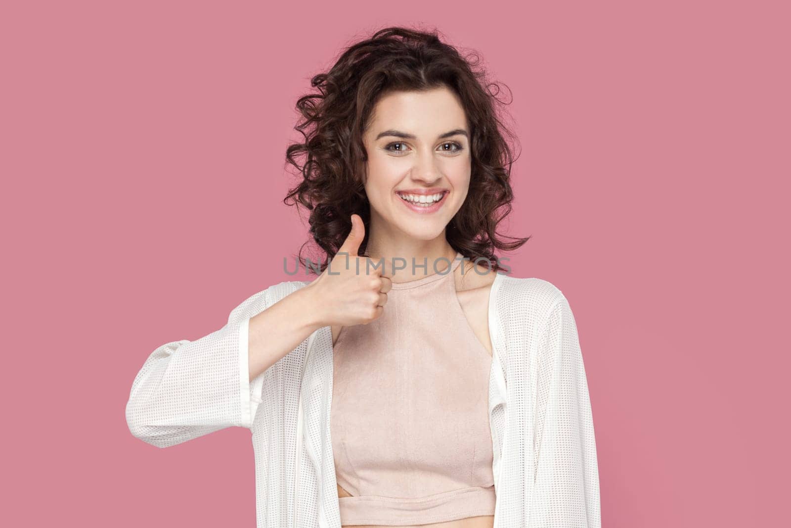 Nice joke. Portrait of woman with curly hairstyle wearing casual style outfit keeps thumb raised, being in good mood, shows her agreement. Indoor studio shot isolated on pink background.