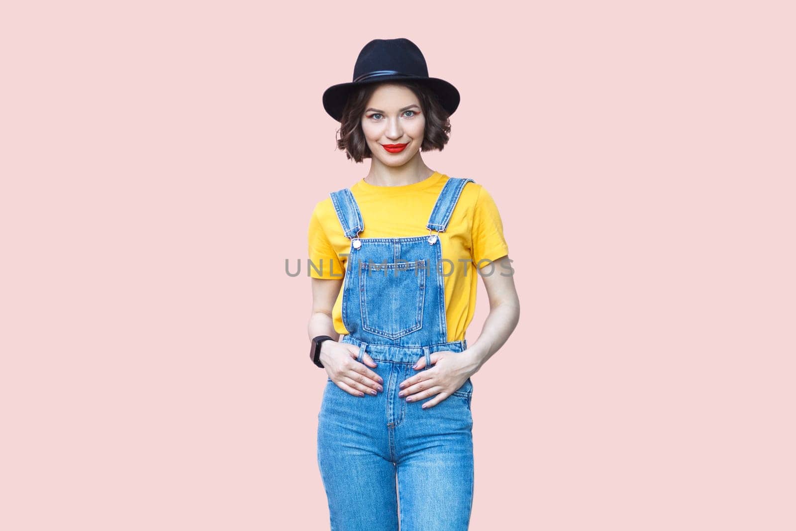 Joyful hipster woman in blue denim overalls looking at camera with happy expression. by Khosro1