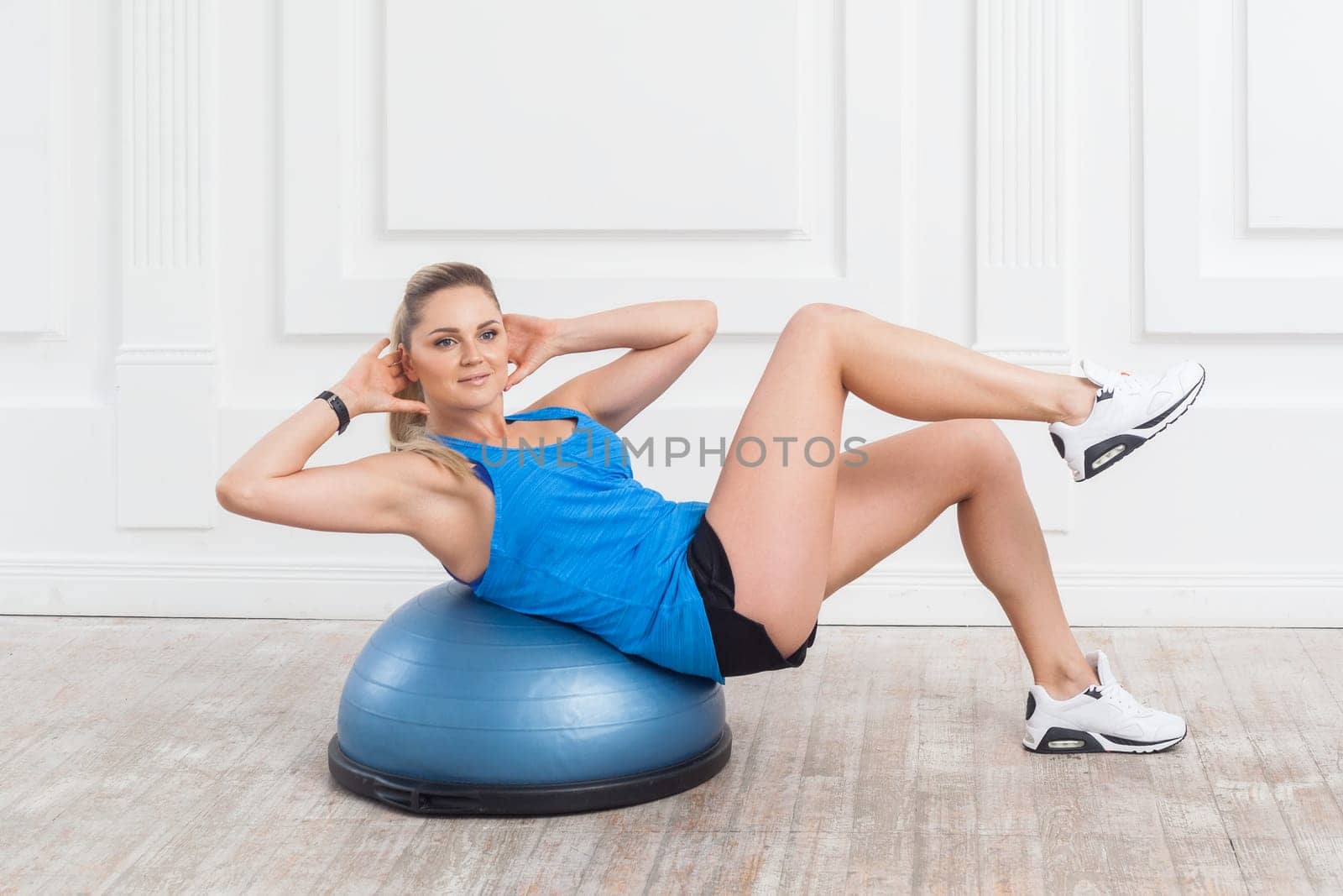 Side view portrait of sporty beautiful blonde woman in black shorts and blue top working in gym doing exercises for abdominal muscles on bosu balance trainer. Indoor studio shot.