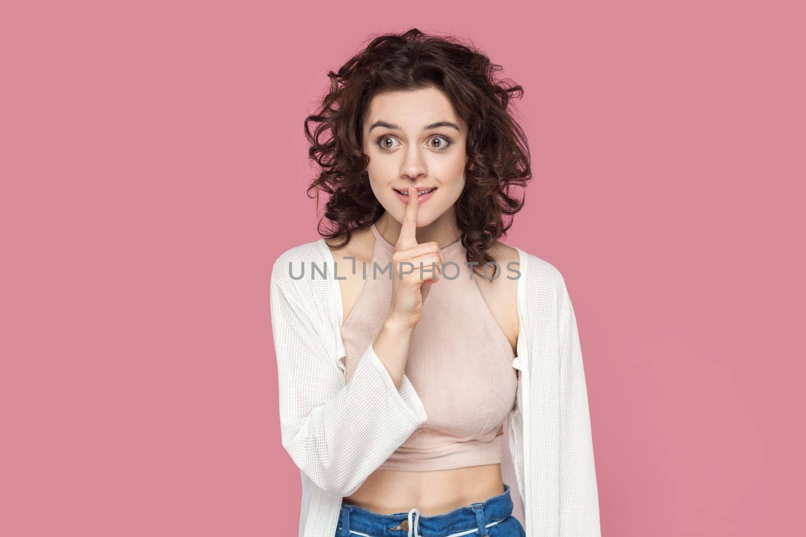 Portrait of positive smiling woman with curly hairstyle wearing casual style outfit showing shh gesture, asking to keep secret. Indoor studio shot isolated on pink background.