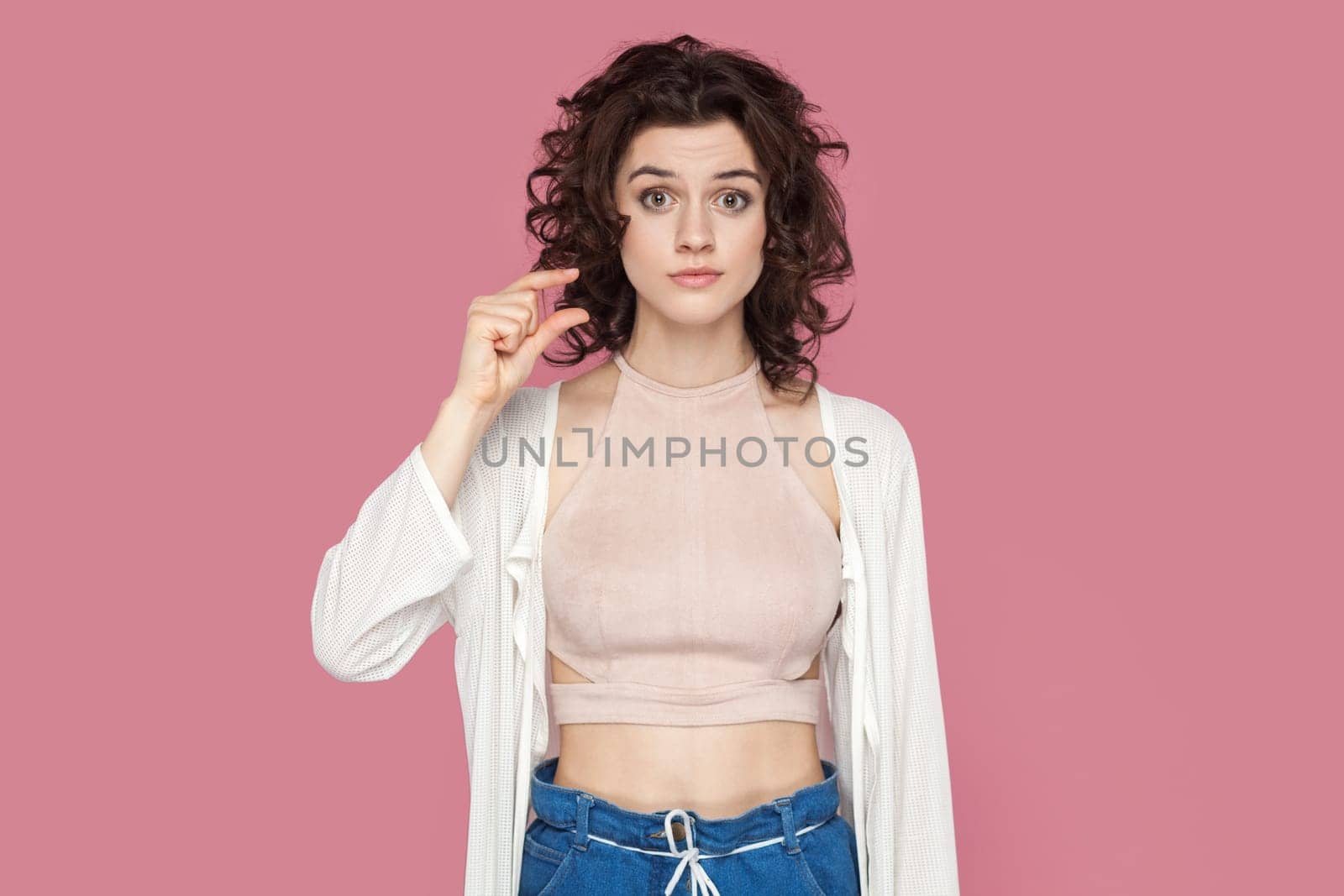 Portrait of confused disappointed beautiful woman with curly hairstyle wearing casual style outfit showing small size with fingers. Indoor studio shot isolated on pink background.