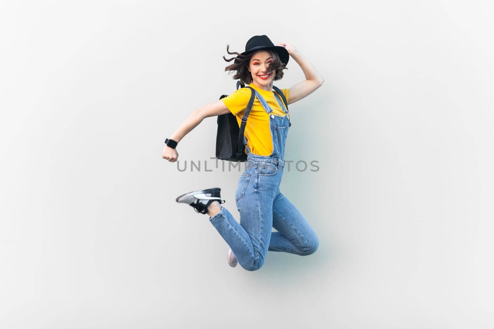 Portrait of attractive overjoyed hipster woman in blue denim overalls, yellow T-shirt and black hat, jumping, being in good mood, expressing happiness. Indoor studio shot isolated on gray background.