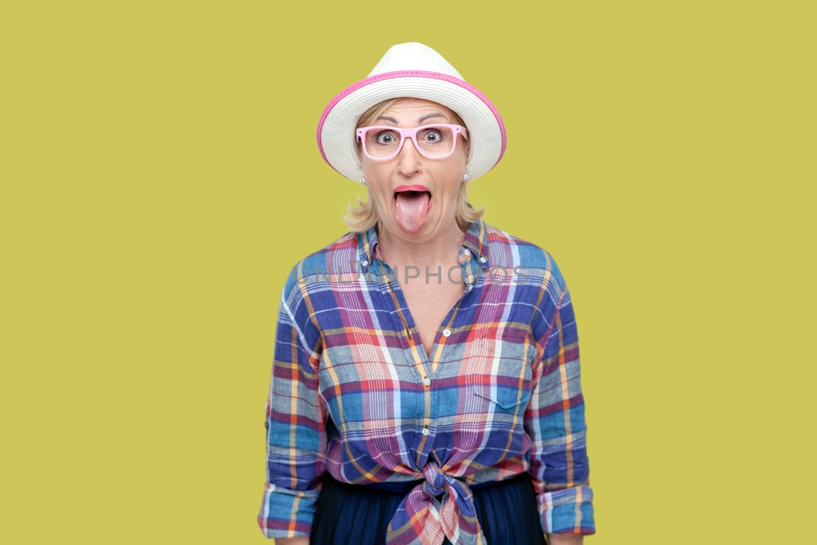 Portrait of childish mature woman wearing checkered shirt, hat and eyeglasses sticking out tongue, making funny grimace. Indoor studio shot isolated on yellow background.