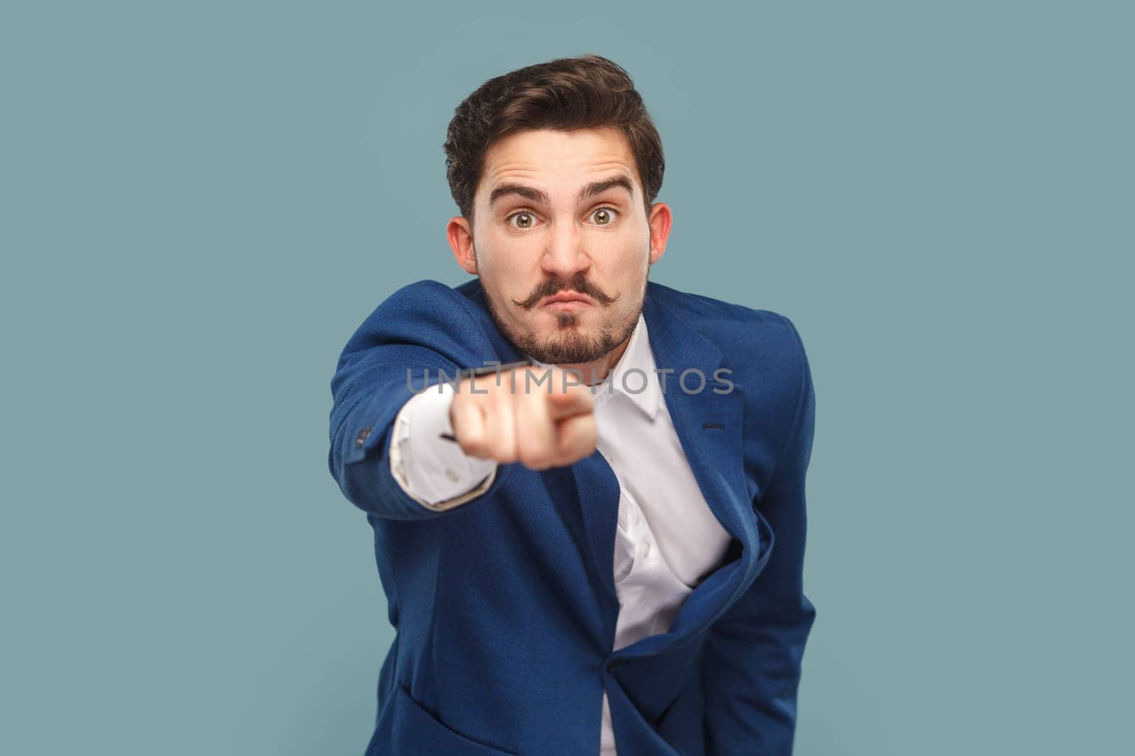 Portrait of serious man with mustache with negative facial expression pointing finger to camera, selecting you, wearing official style suit. Indoor studio shot isolated on light blue background.