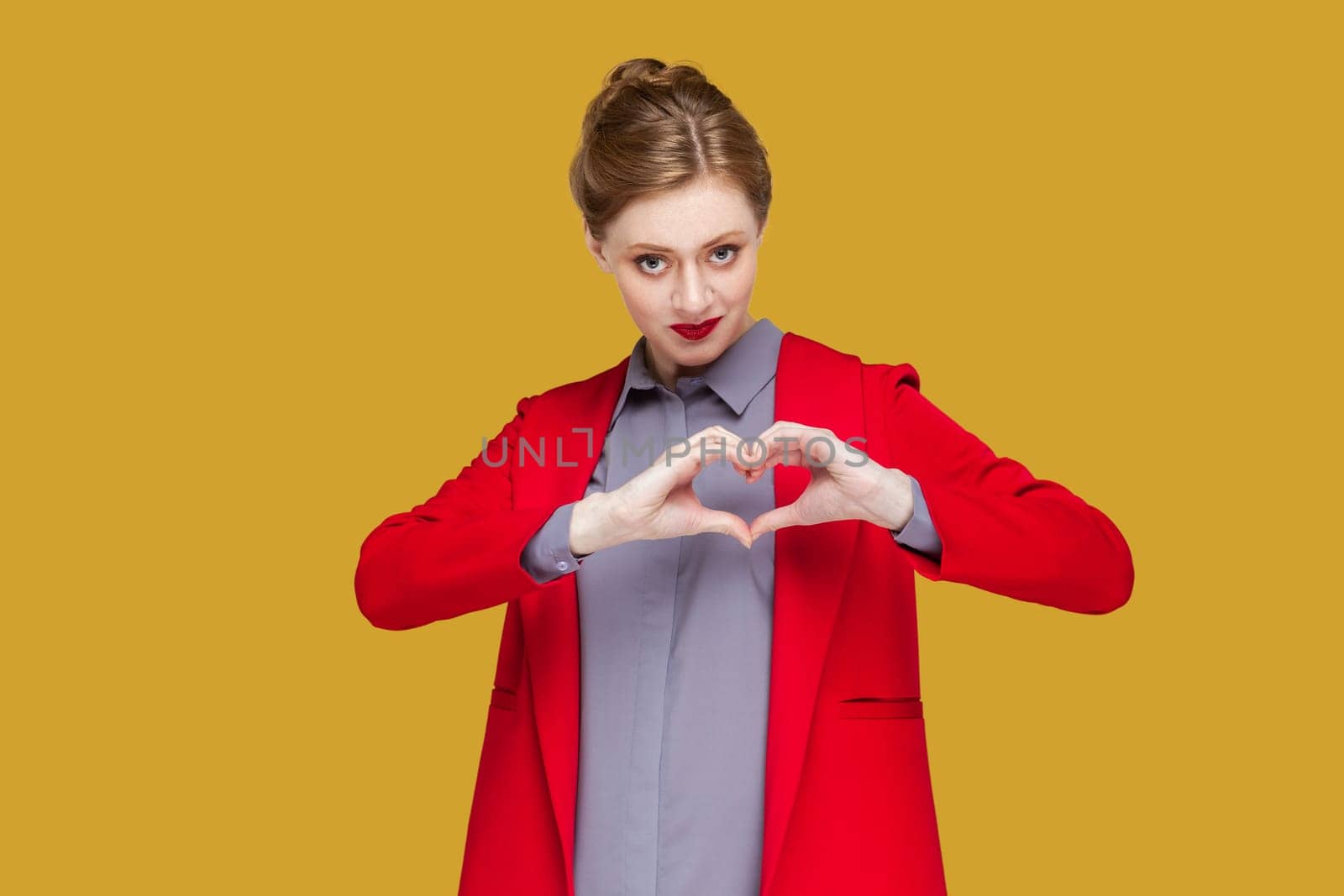 Portrait of romantic flirting woman with red lips standing showing heart shape with hands, looking at camera, expressing devotion, wearing red jacket. Indoor studio shot isolated on yellow background.