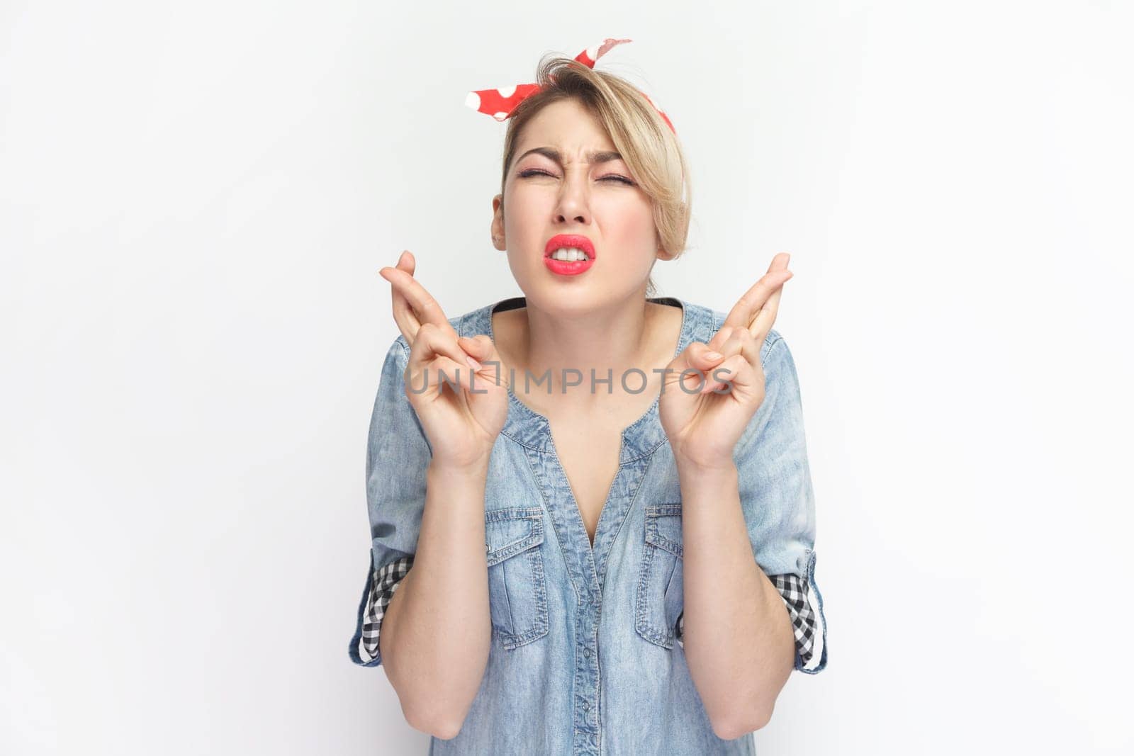 Portrait of hopeful attractive blonde woman wearing blue denim shirt and red headband standing with crossed fingers, praying for good luck. Indoor studio shot isolated on gray background.