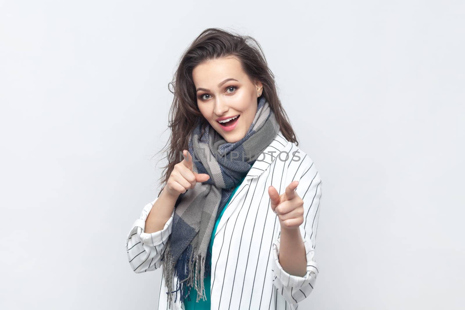 Portrait of overjoyed happy positive brunette woman pointing at camera with index fingers, choosing you, wearing striped jacket and scarf. Indoor studio shot isolated on gray background.