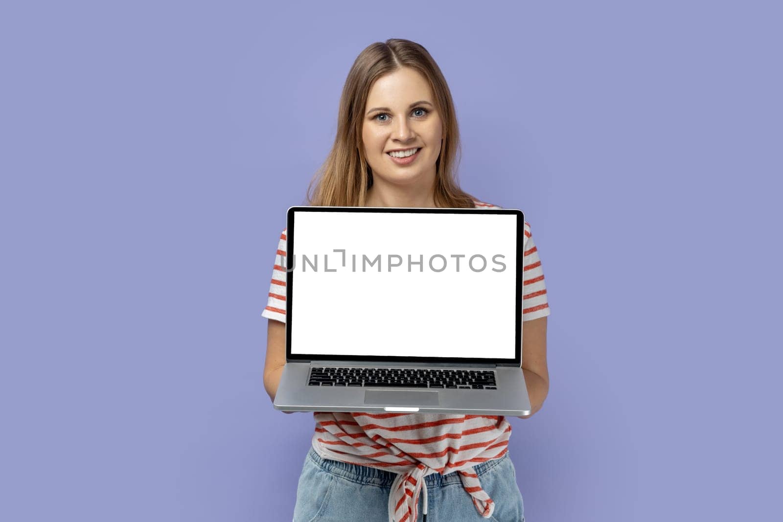 Portrait of happy joyful blond woman wearing striped T-shirt holding laptop with white empty display with empty space for advertisement. Indoor studio shot isolated on purple background.