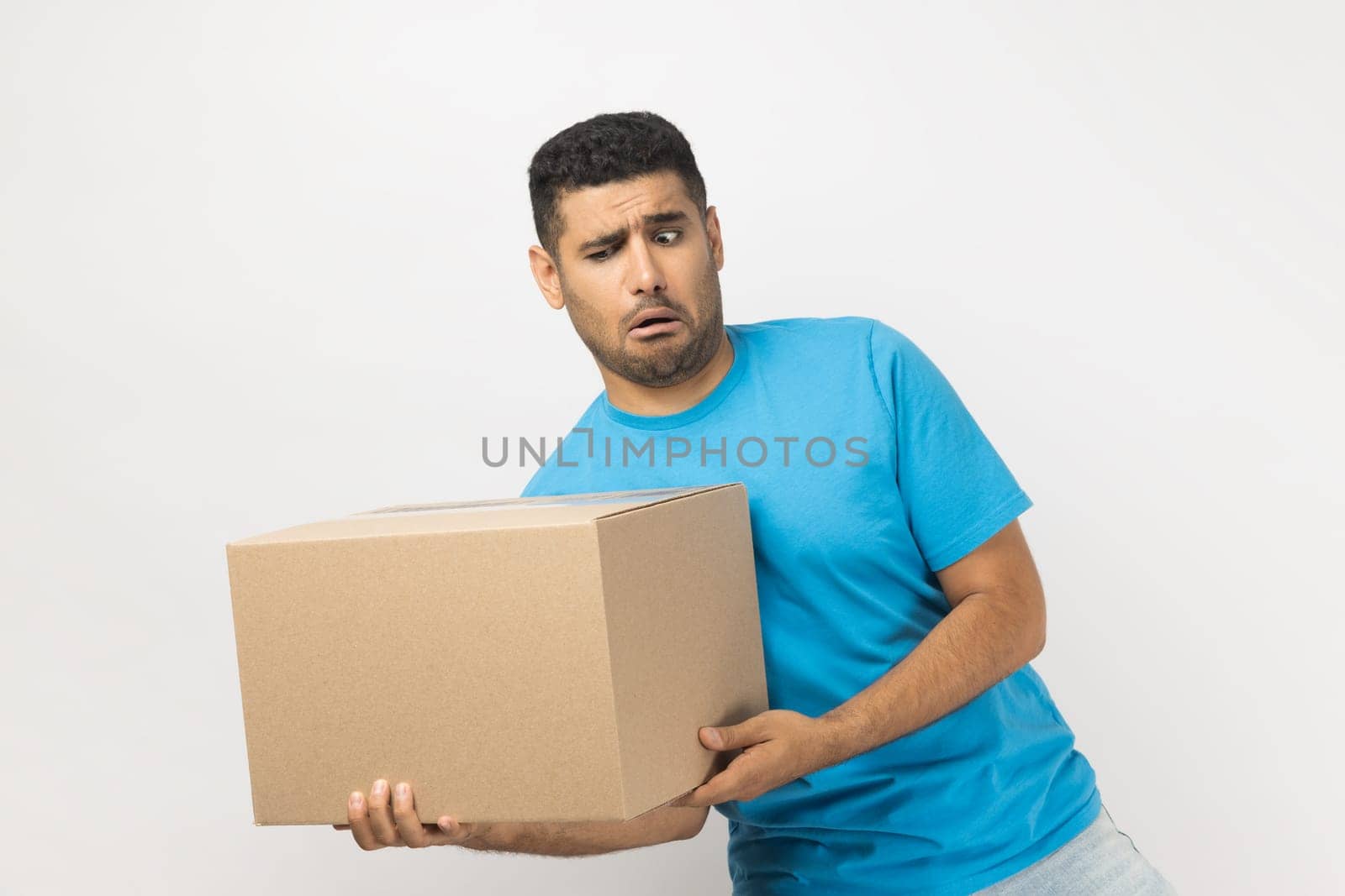 Portrait of funny positive optimistic unshaven man wearing blue T- shirt standing holding very heavy cardboard parcel, making effort to carry box. Indoor studio shot isolated on gray background.